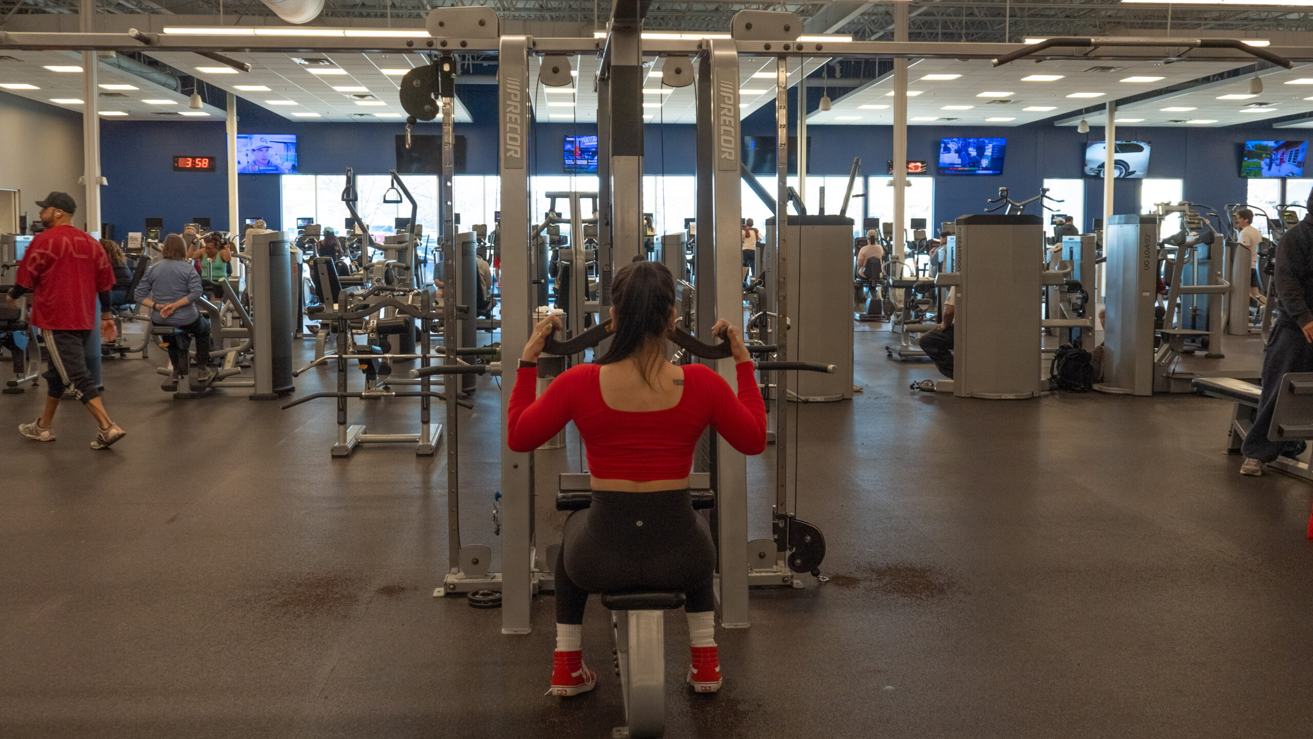 Woman seated at a Precor lat pulldown machine, gripping a straight bar overhead with both hands, photographed from behind in a busy gym.