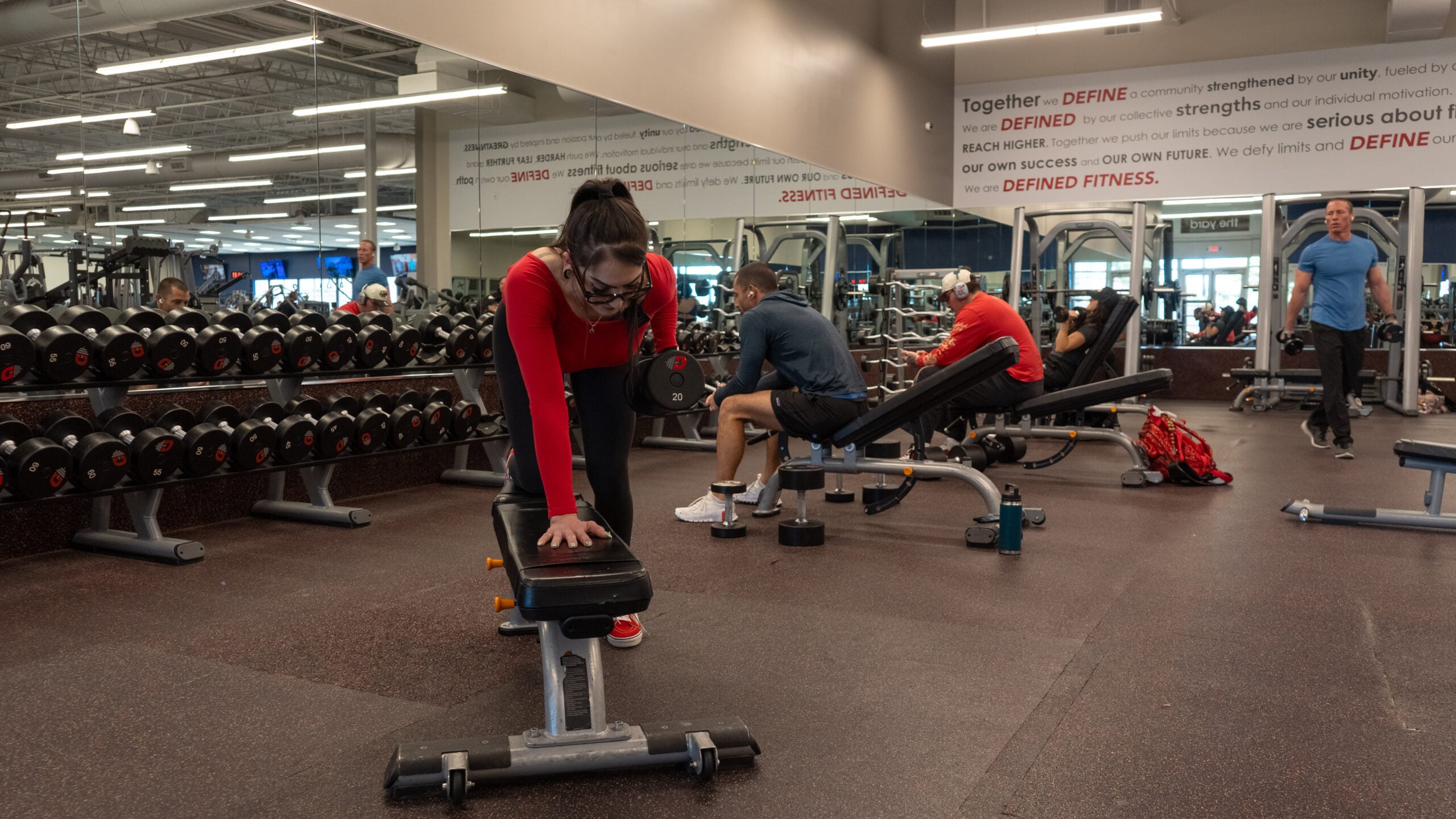 Woman setting up for a single-arm dumbbell row, leaning over a flat bench with one hand braced on the pad, reaching down to pick up a dumbbell from the floor.