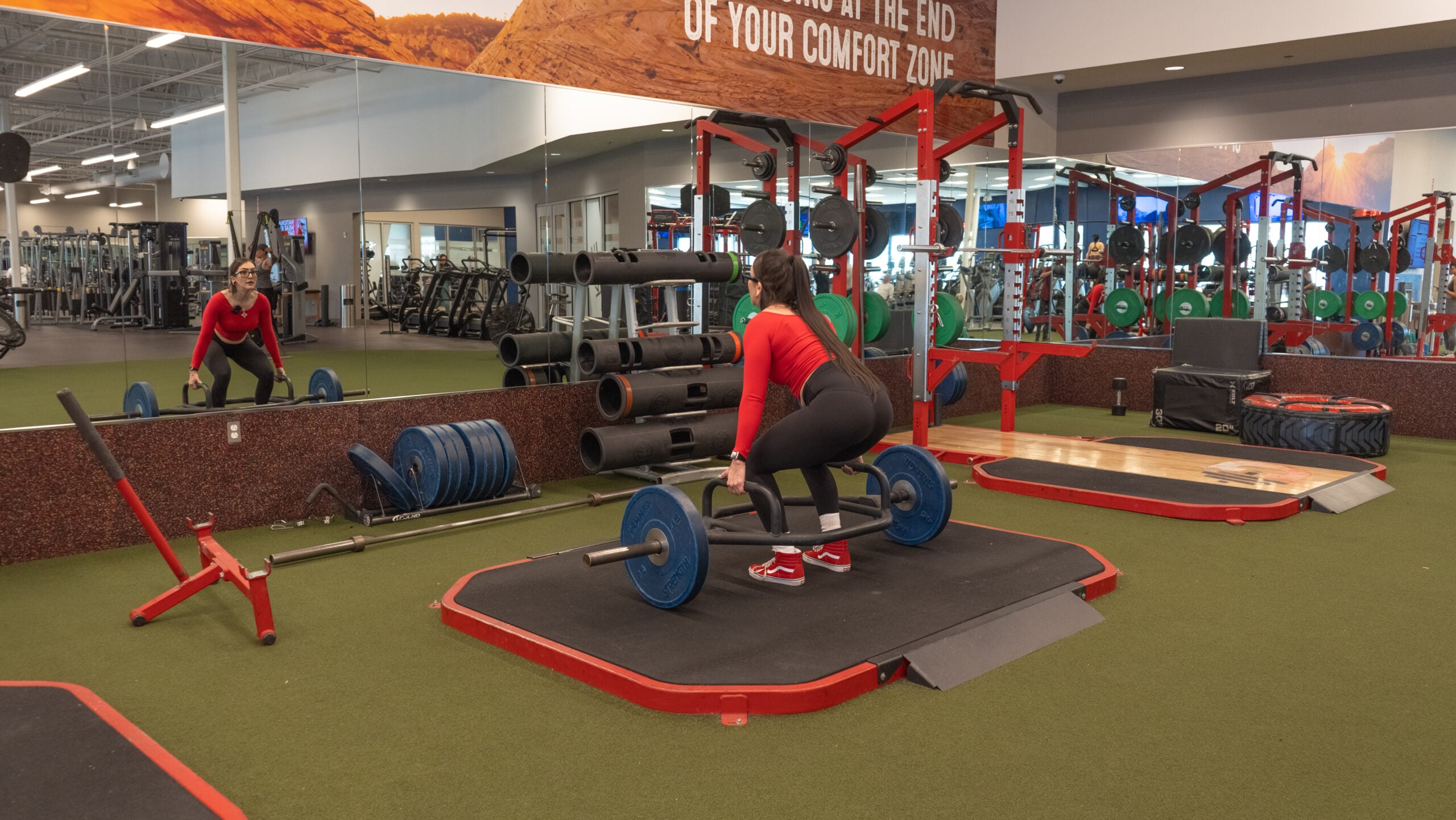 Woman standing upright at the top of a completed trap bar deadlift, holding a loaded hex bar at hip level with arms fully extended, photographed from behind on a lifting platform.
