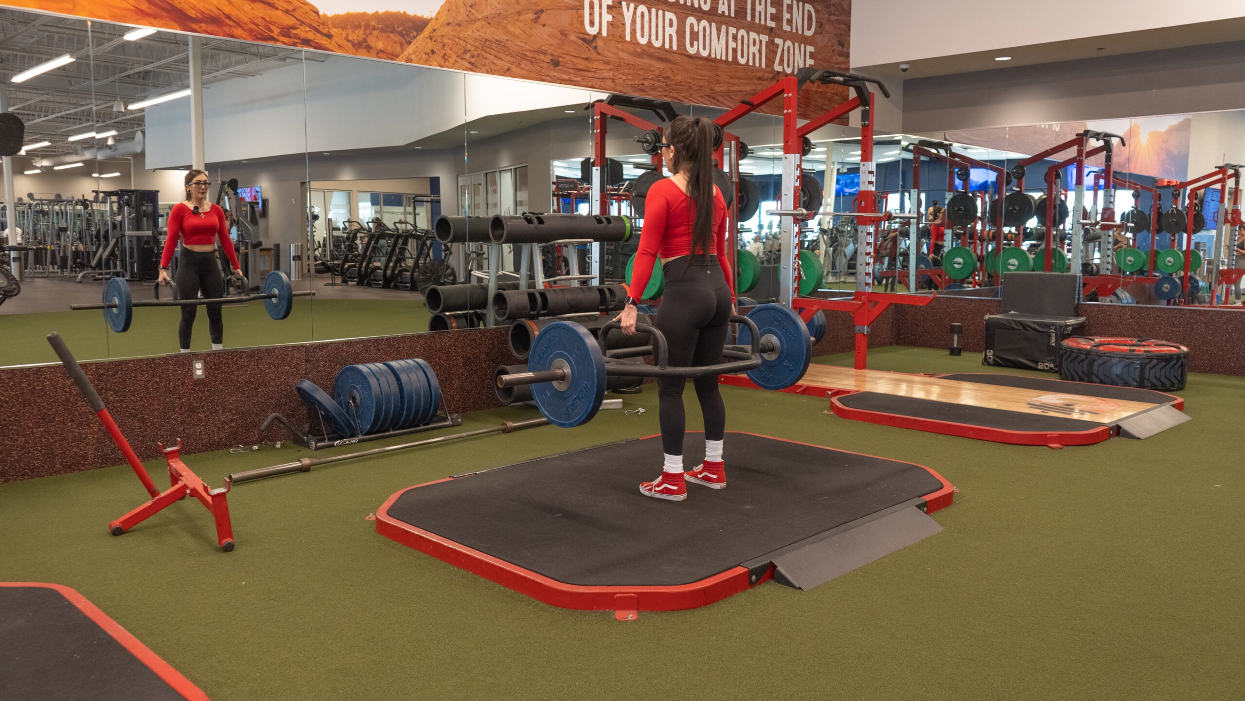 Woman standing at the lockout position of a conventional barbell deadlift, holding a loaded barbell at hip height with arms fully extended, photographed from behind on a lifting platform.