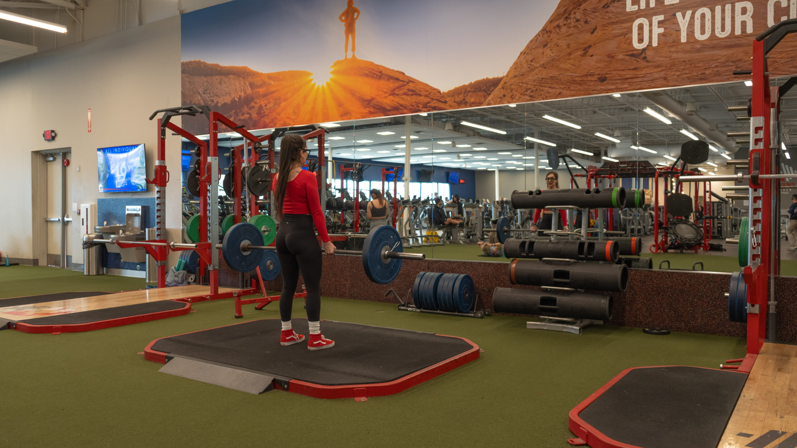 Woman at the setup position of a conventional barbell deadlift, bent forward at the hips with both hands gripping a loaded barbell just above the floor, knees slightly bent and back flat.