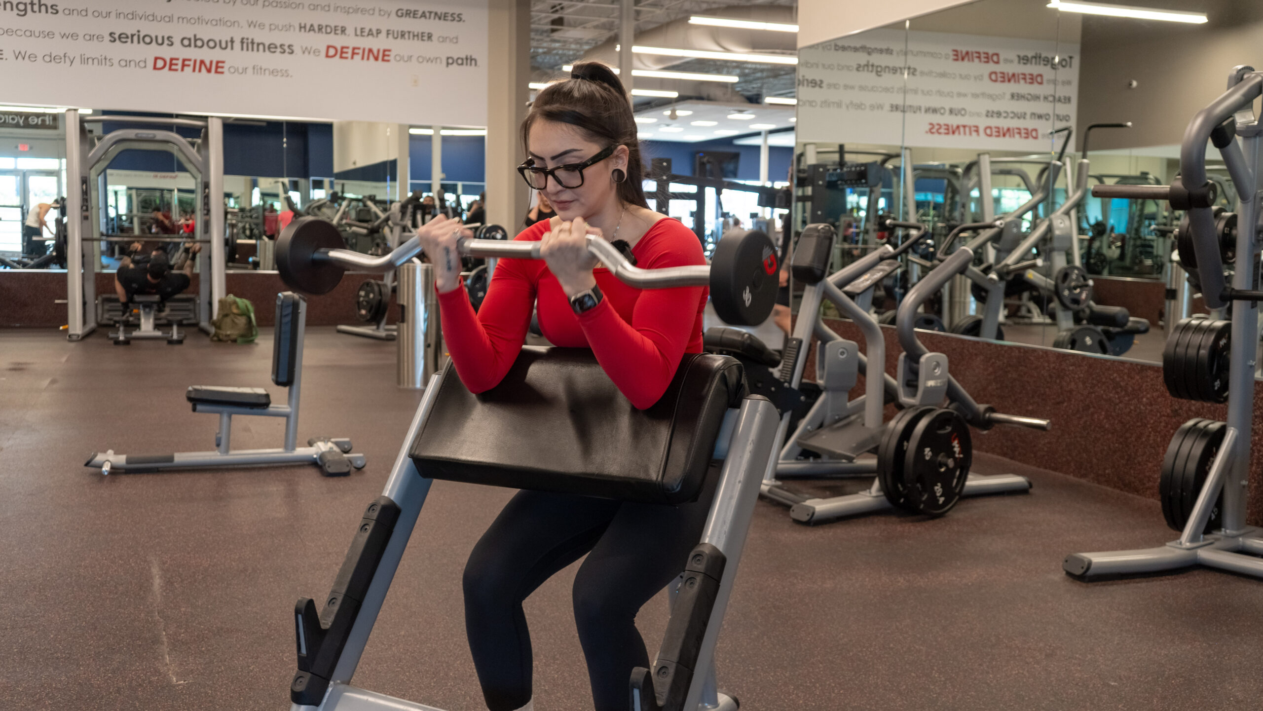Woman performing a preacher curl on a preacher curl bench, arms braced against the padded support, curling an EZ curl bar upward toward her chest at the top of the movement.