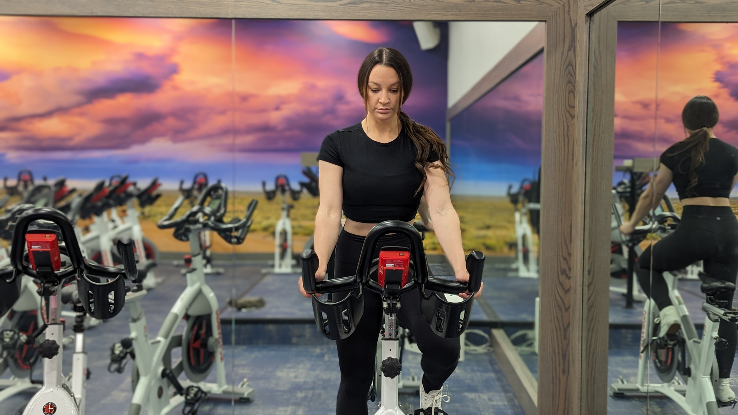 Woman riding a stationary indoor cycling bike in a spin studio, leaning slightly forward over the handlebars, photographed from the front with mirrored walls and additional bikes visible in the background.