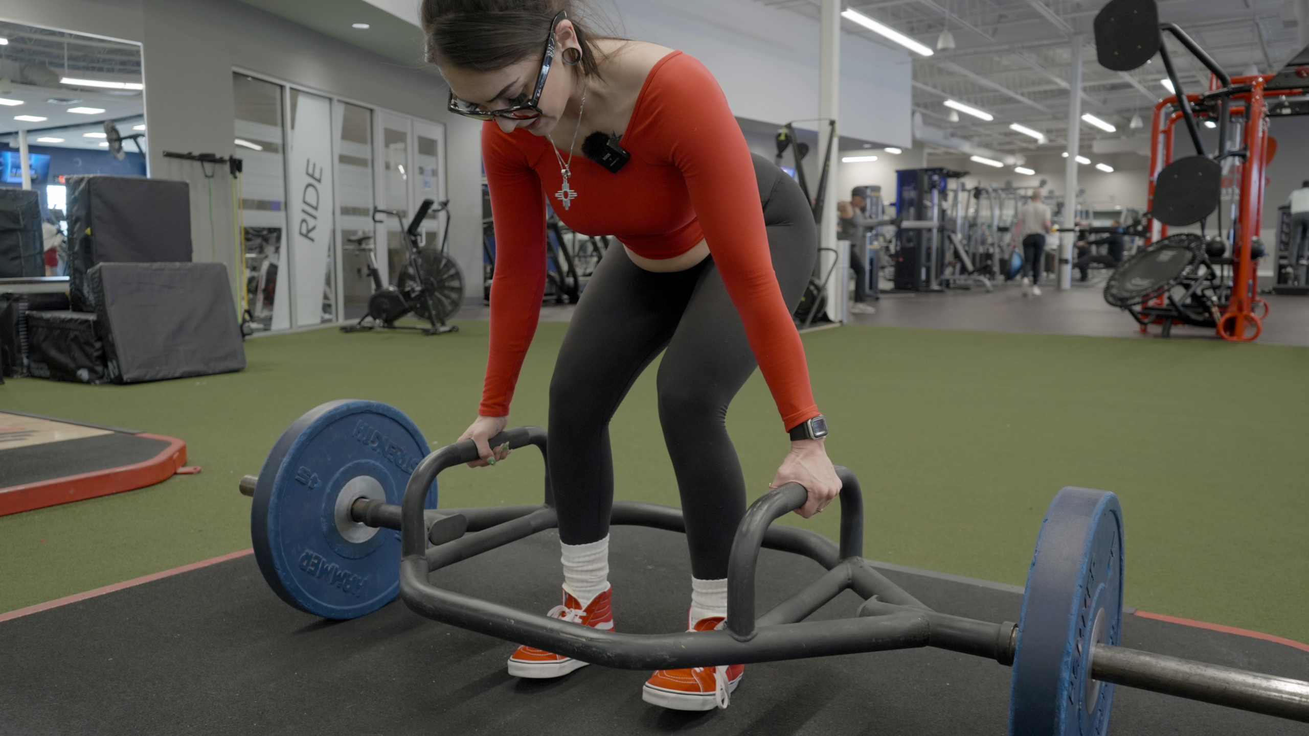 Woman setting up for a trap bar deadlift, leaning forward to grip the handles of a loaded hex bar on the floor, with weight plates loaded on both sides.